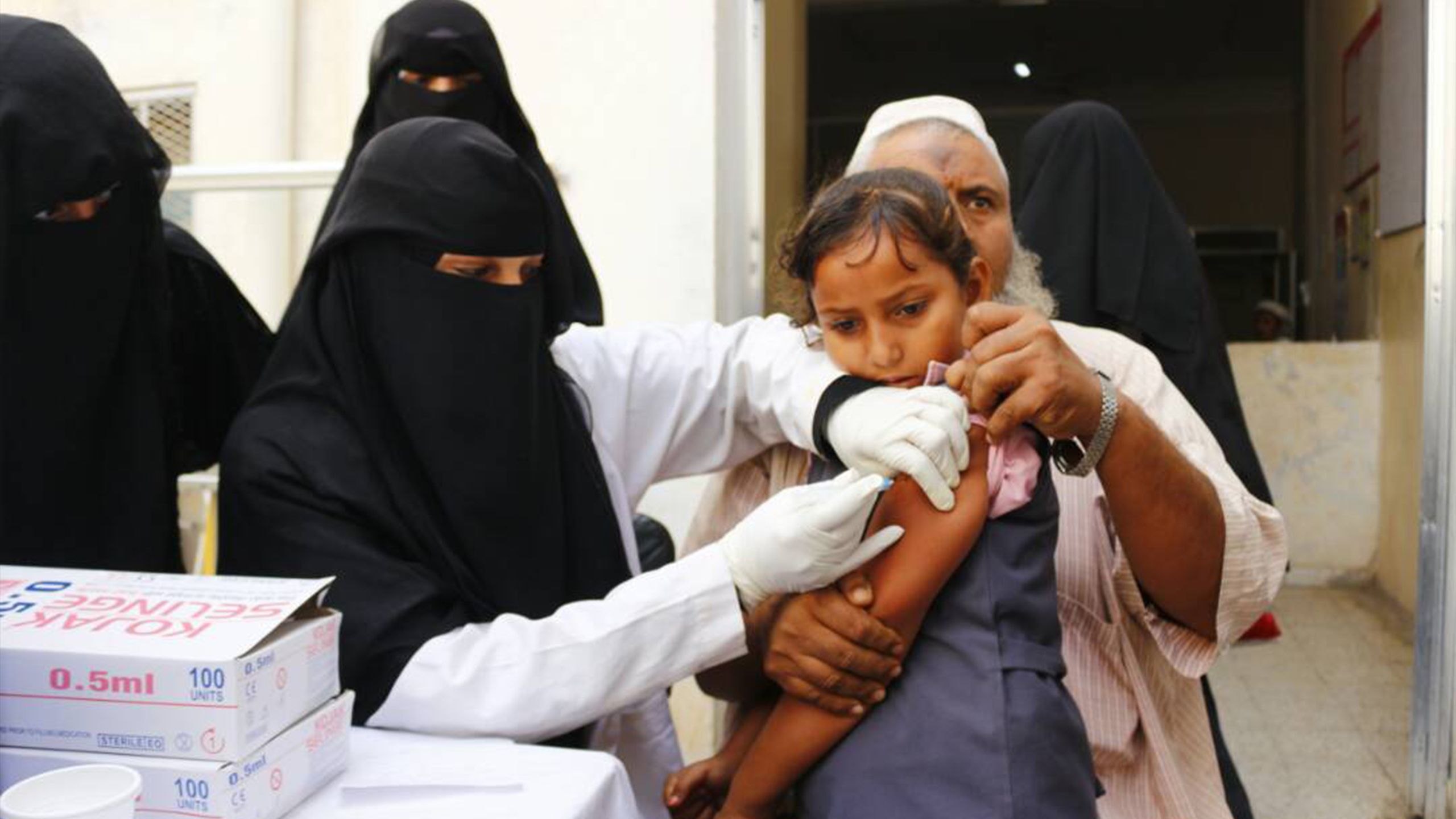 A father holds his daughter as nurses administer a vaccination against diphtheria. Image by Save the Children, alert 200 Yemen.